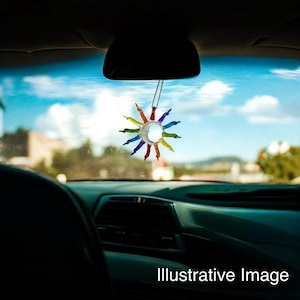 Puede incluir: Un amuleto de coche con forma de sol de colores arcoíris que cuelga del espejo retrovisor de un coche. El amuleto está hecho de vidrio y tiene un centro transparente.
