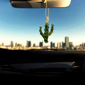 May include: A green glass cactus car charm hanging from a gold chain, suspended from a car's rearview mirror. The charm is in focus against a blurred cityscape and a clear blue sky.