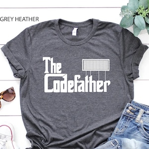 May include: A dark grey heather t-shirt with the text "The Codefather" in white, featuring a stylized image of a computer circuit board.