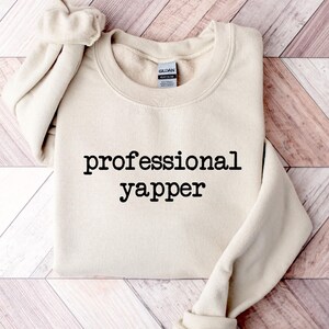 May include: A beige sweatshirt with the text "professional yapper" printed in black on the front.