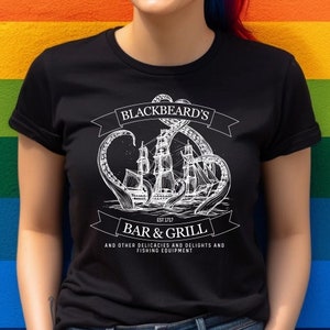 May include: A black t-shirt with a white graphic of a ship being attacked by a giant octopus. The text on the shirt reads "Blackbeard's Est. 1973 Bar & Grill and other delicacies and delights and fishing equipment."