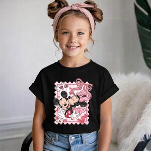 Puede incluir: Camiseta negra con un gr&aacute;fico de Mickey y Minnie Mouse abraz&aacute;ndose dentro de un dise&ntilde;o de sello postal rosa. El dise&ntilde;o incluye corazones y un lazo rosa. La camiseta la lleva una ni&ntilde;a con una diadema rosa.