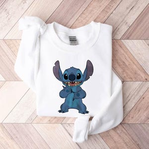 Pode incluir: Camisola branca com um personagem de desenho animado azul com orelhas grandes e um sorriso largo. O personagem levanta a m&atilde;o em um gesto. A camisola &eacute; feita de um material macio e &eacute; adequada para uso casual.