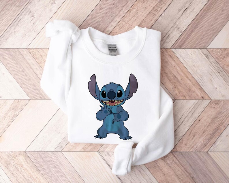 Pode incluir: Camisola branca com um personagem de desenho animado azul com orelhas grandes e um sorriso largo. O personagem levanta a m&atilde;o em um gesto. A camisola &eacute; feita de um material macio e &eacute; adequada para uso casual.