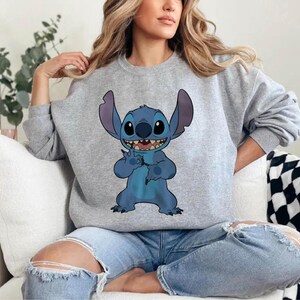 Pode incluir: Uma camisola cinza claro com um personagem de desenho animado azul com orelhas grandes e um sorriso largo. O personagem est&aacute; levantando dois dedos. A camisola &eacute; usada com jeans azuis.