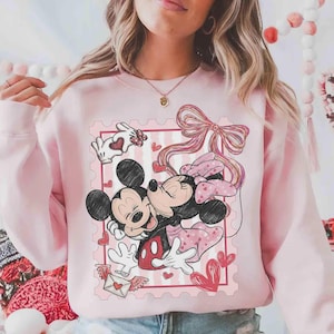 Puede incluir: Sudadera rosa claro con un dise&ntilde;o de Mickey y Minnie Mouse abraz&aacute;ndose dentro de un dise&ntilde;o de sello postal rosa. El dise&ntilde;o incluye corazones, un lazo y una carta de amor. La sudadera es de color rosa claro liso.