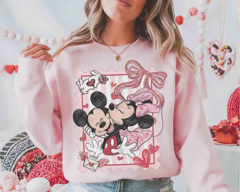 Puede incluir: Sudadera rosa claro con un dise&ntilde;o de Mickey y Minnie Mouse abraz&aacute;ndose dentro de un dise&ntilde;o de sello postal rosa. El dise&ntilde;o incluye corazones, un lazo y una carta de amor. La sudadera es de color rosa claro liso.