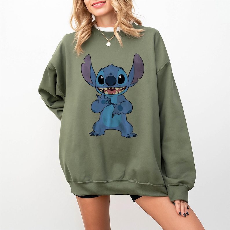 Pode incluir: Camisola verde-oliva com um personagem de desenho animado azul com orelhas grandes e um sorriso largo. O personagem est&aacute; segurando um pequeno objeto. A camisola &eacute; de estilo casual, adequada para o uso di&aacute;rio.