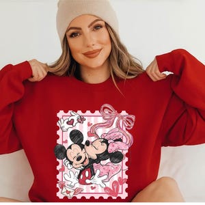 Puede incluir: Una sudadera roja con un gr&aacute;fico de Mickey y Minnie Mouse abraz&aacute;ndose dentro de un dise&ntilde;o de sello postal rosa. El dise&ntilde;o incluye corazones y un lazo rosa. La sudadera es de color rojo liso.