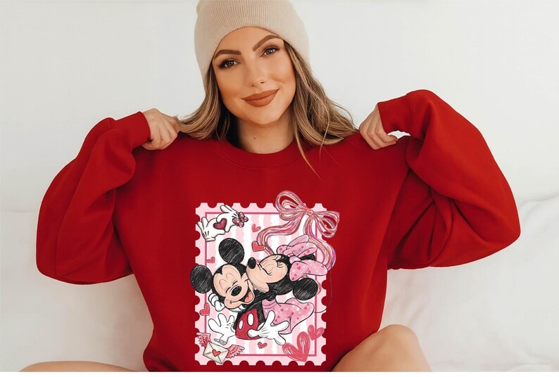 Puede incluir: Una sudadera roja con un gr&aacute;fico de Mickey y Minnie Mouse abraz&aacute;ndose dentro de un dise&ntilde;o de sello postal rosa. El dise&ntilde;o incluye corazones y un lazo rosa. La sudadera es de color rojo liso.