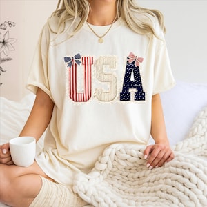 May include: Cream-colored t-shirt with the word "USA" in patriotic colors. The "U" is red and white striped, the "S" is beige, and the "A" is navy blue with white stars. Each letter has a decorative bow.
