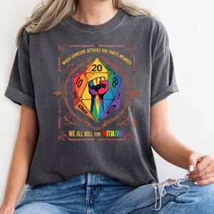 May include: Dark gray t-shirt featuring a colorful graphic of a D20 die with a rainbow fist. The text reads "WHEN SOMEONE ATTACKS ONE PARTY MEMBER" and "WE ALL ROLL FOR INITIATIVE". The shirt is a casual wear item.