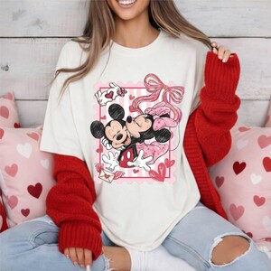 Puede incluir: Camiseta color crema con un gr&aacute;fico de Mickey y Minnie Mouse abraz&aacute;ndose, dentro de un dise&ntilde;o de sello postal rosa con corazones y un lazo. Las mangas son rojas.
