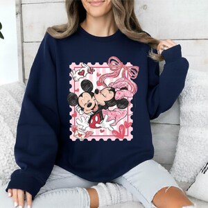 Puede incluir: Sudadera azul marino con un dise&ntilde;o de Mickey y Minnie Mouse abraz&aacute;ndose dentro de un dise&ntilde;o de sello postal rosa. El dise&ntilde;o incluye corazones, lazos y una carta de amor, creando un tema rom&aacute;ntico. La sudadera es una prenda c&oacute;moda e informal.