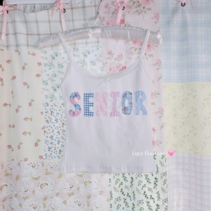 May include: White camisole top with spaghetti straps and lace detailing. The word "SENIOR" is appliquéd across the front in floral and gingham fabrics. The top is displayed on a white hanger, set against a backdrop of floral and patterned fabrics.