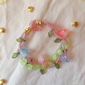 Op de afbeelding: Een handgemaakte armband met roze kralen, versierd met doorschijnende roze, blauwe en groene bloem- en bladbedels. De armband heeft ook kleine groene kralen met de letters "FOR" en "L". Gouden stervormige versieringen zijn rond de armband verspreid.