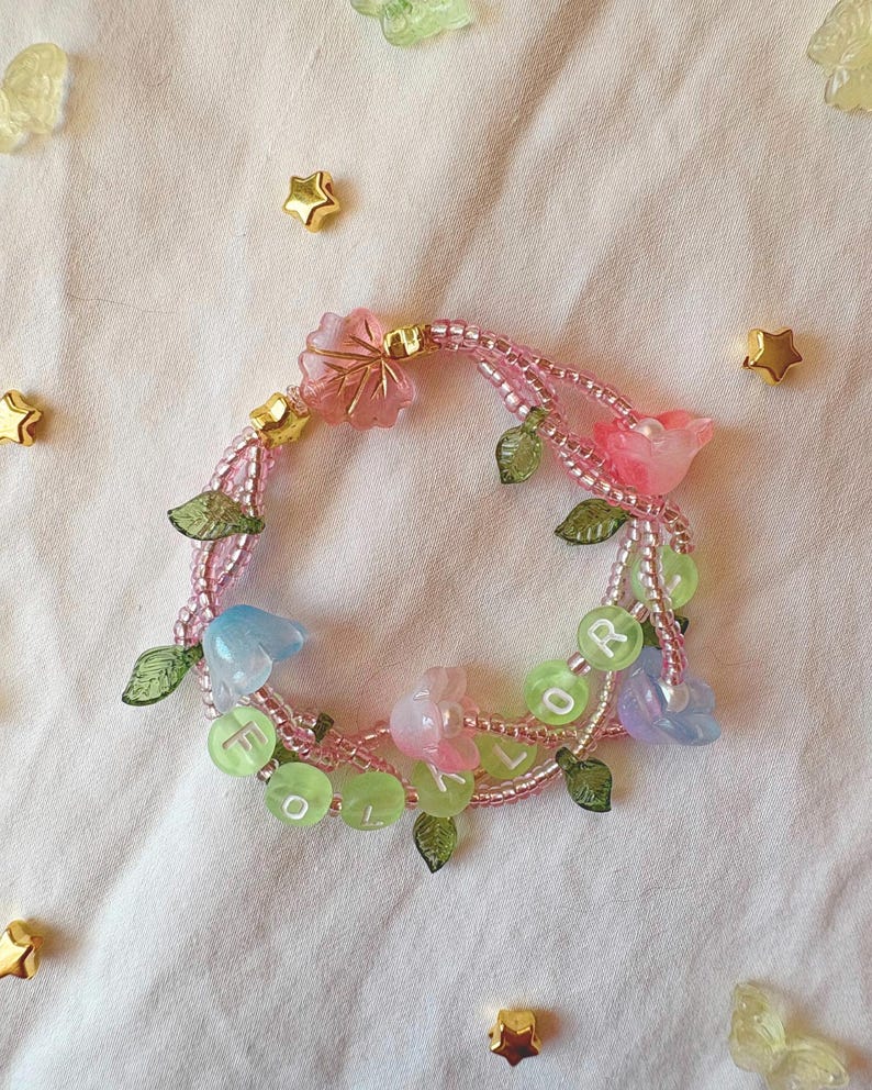 Op de afbeelding: Een handgemaakte armband met roze kralen, versierd met doorschijnende roze, blauwe en groene bloem- en bladbedels. De armband heeft ook kleine groene kralen met de letters "FOR" en "L". Gouden stervormige versieringen zijn rond de armband verspreid.