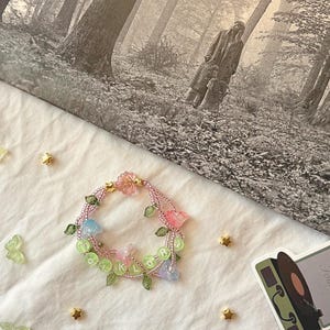 Op de afbeelding: Een kralenarmband met roze, blauwe en heldere bloemaccenten en het woord "folklore" in groene kralen. De armband ligt op een wit oppervlak met gouden ster- en vlinderdecoraties.