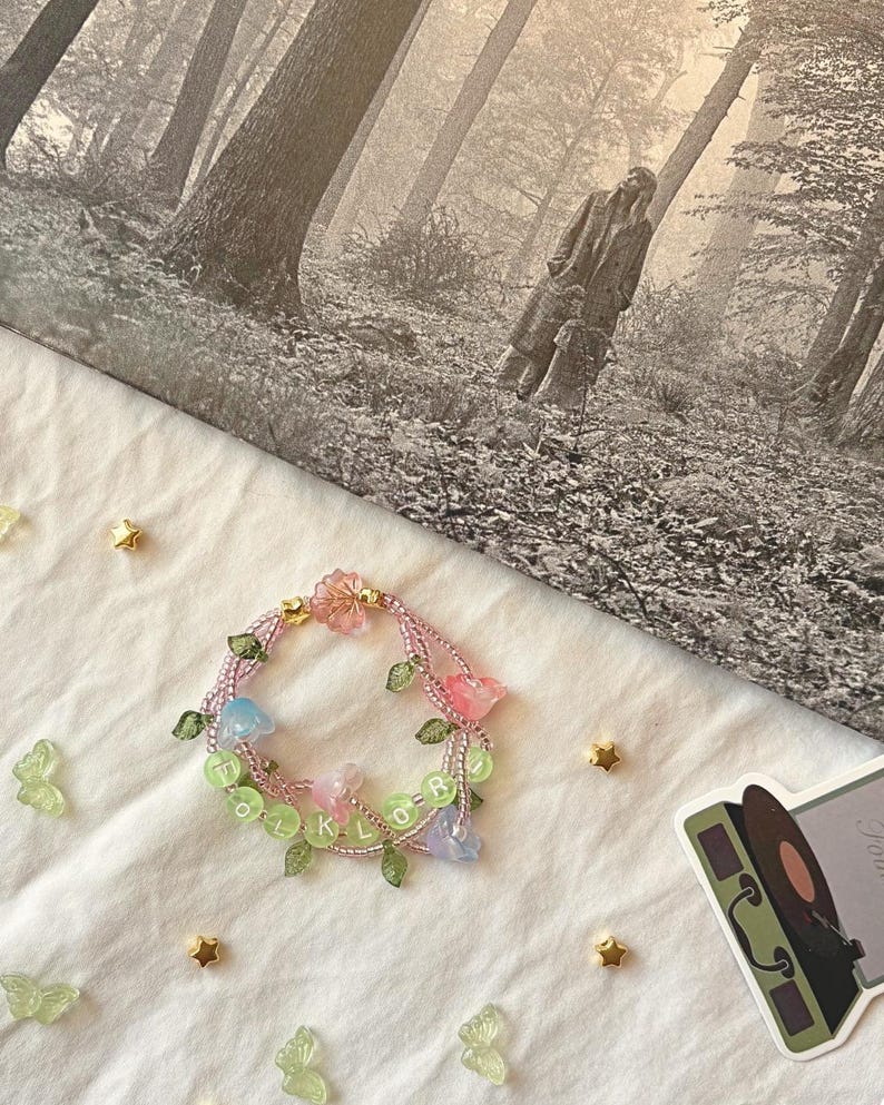 Op de afbeelding: Een kralenarmband met roze, blauwe en heldere bloemaccenten en het woord "folklore" in groene kralen. De armband ligt op een wit oppervlak met gouden ster- en vlinderdecoraties.