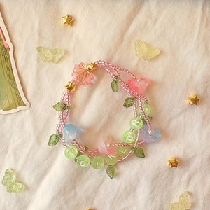 Op de afbeelding: Een handgemaakte armband met roze en gouden kralen, versierd met roze en blauwe bloemvormige bedels, groene bladaccenten en groene letterkralen met de tekst "FLOR". Gouden sterren en doorschijnende vlinders zijn verspreid.