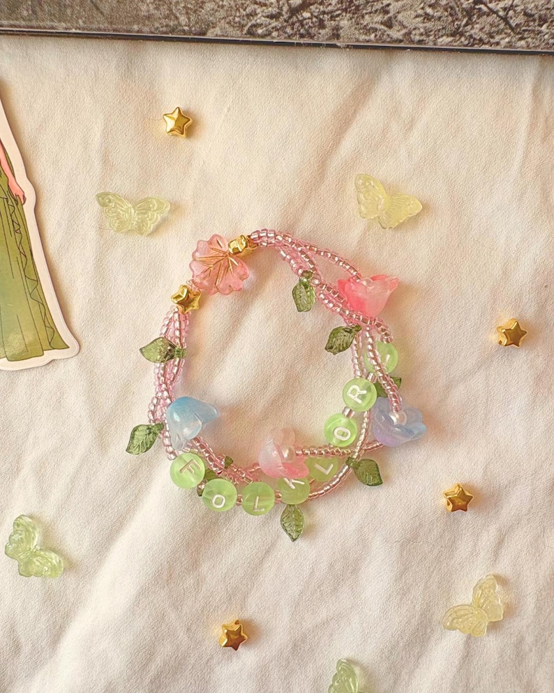 Op de afbeelding: Een handgemaakte armband met roze en gouden kralen, versierd met roze en blauwe bloemvormige bedels, groene bladaccenten en groene letterkralen met de tekst "FLOR". Gouden sterren en doorschijnende vlinders zijn verspreid.
