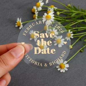 May include: A clear, round sticker with gold lettering that reads "Save the Date" with the date "February 28, 2026" and names "Vittoria & Richard". The sticker is held in front of a background of daisies.