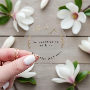 Op de afbeelding: Een heldere, ronde sticker met gouden letters en een gouden bladontwerp. De tekst luidt "Thank you for celebrating with us" en de namen "Mr + Mrs Anderson". De sticker wordt door een hand vastgehouden, omringd door witte bloemen en groene bladeren.
