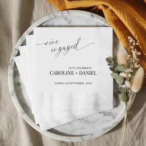 May include: White paper napkins with the text "we're engaged" in elegant script, along with "Let's Celebrate" and the names Caroline + Daniel, and the date Sunday 20 September, 2026. The napkins are on a marble tray.