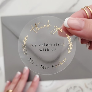 Op de afbeelding: Transparante, ronde sticker met gouden belettering. De tekst luidt "Thank you", "for celebrating with us" en "Mr + Mrs Parker". Voorzien van een decoratieve bladerenrand. Ideaal voor huwelijksbedankjes.