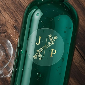 Puede incluir: Primer plano de una botella de vidrio verde con una etiqueta circular transparente. La etiqueta presenta las letras doradas "J" y "P" con detalles florales. Se ven gotas de agua en la superficie de la botella.