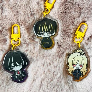 Puede incluir: Tres llaveros de personajes de estilo anime con cierres amarillos. Cada llavero presenta un diseño de personaje diferente con contornos negros y varios colores de cabello. Los llaveros se muestran sobre una superficie rosa y esponjosa.