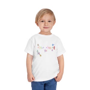 May include: A white toddler t-shirt with the words "First Timer" in colorful, handwritten lettering. The shirt features several colorful star and kite-like drawings. The child is wearing blue pants.