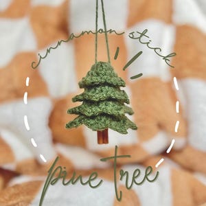 Peut inclure: Une décoration de sapin de Noël en crochet vert suspendue à un bâton de cannelle. L'ornement est suspendu sur un fond à carreaux brun et blanc. Le texte "cinnamon stick" et "pine tree" est écrit en cursive sur l'image.