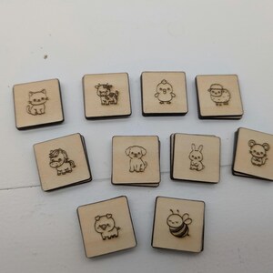Könnte beinhalten: Eine Sammlung von zehn kleinen, quadratischen Holzfliesen mit lasergravierten Tierillustrationen. Die Tiere umfassen eine Katze, eine Kuh, ein Küken, ein Schaf, ein Pferd, einen Hund, ein Kaninchen, eine Maus, ein Schwein und eine Biene. Jede Fliese hat einen dunkelbraunen Rand.