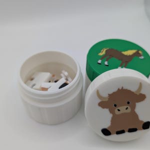 May include: A white plastic container with a lid featuring a brown Highland cow illustration. A green lid with a brown horse illustration is also visible. The container is open, revealing small, white objects inside.