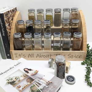 May include: A wooden spice rack with glass jars filled with various spices. The jars have black labels with the names of the spices written on them. The rack is labelled "It's All About The Spice".