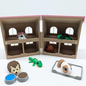 Op de afbeelding: Een miniatuur poppenhuis met een roze dak en verschillende kleine dierenbeeldjes. De set bevat een egel, een gekko, een cavia en een vis. Er zijn ook twee kommen en een klein bed.