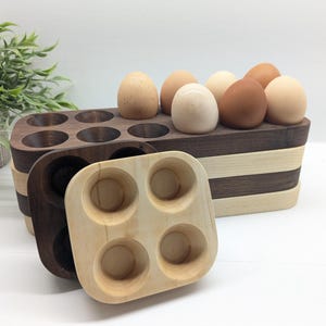 Könnte beinhalten: Eierhalter aus Holz in verschiedenen Braun- und Naturholztönen, die braune und weiße Eier halten. Ein kleiner Halter für vier Eier befindet sich im Vordergrund. Die Eierhalter stehen auf einer weißen Oberfläche, mit einer Topfpflanze im Hintergrund.