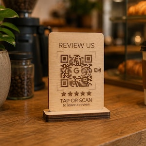 Peut inclure: Un panneau en bois avec le texte "REVIEW US" et un code QR, le logo Google et le symbole NFC. En dessous du code QR se trouvent cinq étoiles et le texte "TAP OR SCAN to leave a review". Le panneau est posé sur une surface en bois.
