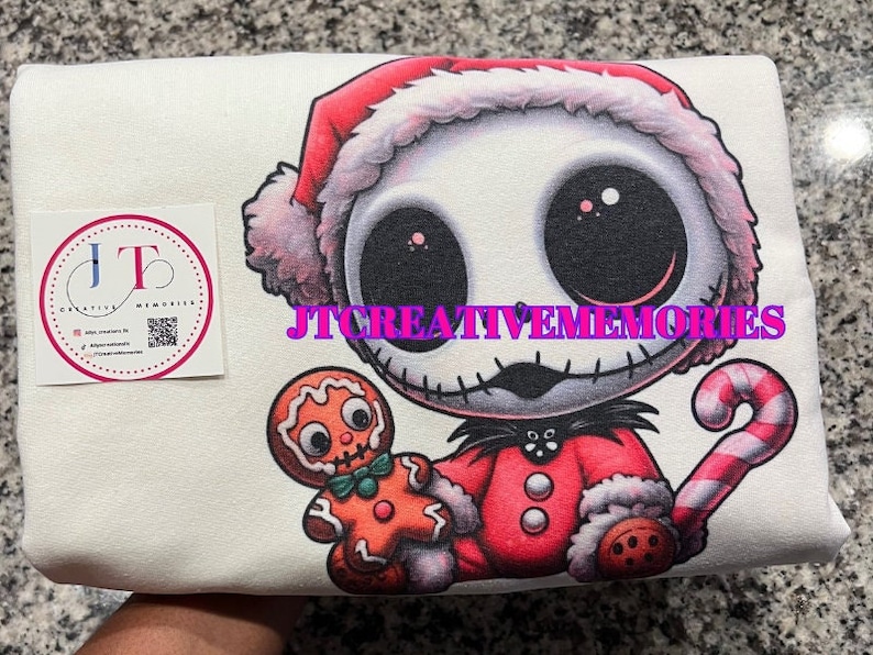May include: White t-shirt with a cartoon image of Jack Skellington from The Nightmare Before Christmas dressed as Santa Claus. He is holding a gingerbread man and a candy cane. The text "JT CREATIVE MEMORIES" is printed on the shirt.