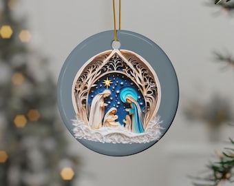 Puede incluir: Un adorno redondo de cerámica con una escena de la natividad azul y blanca. La escena está rodeada por un diseño de filigrana dorada y un cielo estrellado.