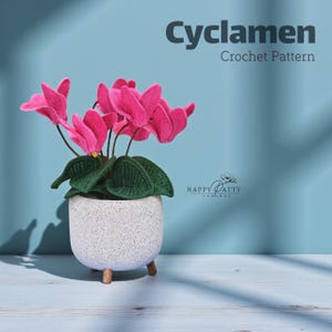 May include: A crochet cyclamen plant in a speckled white pot with wooden legs. The plant features vibrant pink flowers and green leaves. The text "Cyclamen Crochet Pattern" is displayed above the plant.