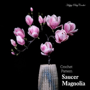 May include: A crocheted saucer magnolia flower arrangement in a blue and white ceramic vase. The flowers are pink and white and have a delicate, realistic look. The text "Crochet Pattern Saucer Magnolia" is displayed below the vase.