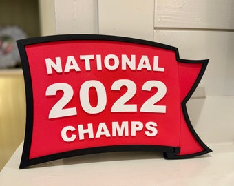 Puede incluir: Un letrero rojo con las palabras "NATIONAL 2022 CHAMPS" en letras blancas. El letrero tiene un borde negro y forma de bandera, ideal para celebrar un logro.
