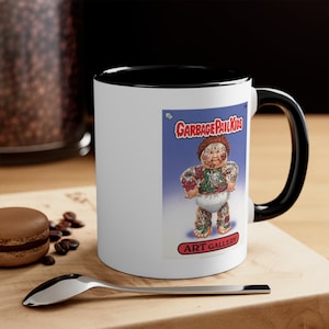 Puede incluir: Taza de cerámica blanca con interior y asa negros. La taza presenta una colorida ilustración de Garbage Pail Kids sobre un fondo azul, con los textos "GARBAGE PAIL KIDS" y "ART GALLERY". Una cuchara y un macaron están sobre la superficie de madera.