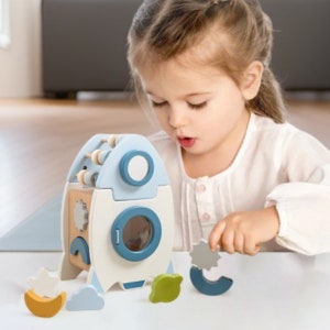 May include: A wooden rocket toy with a blue and white color scheme. The rocket has a small abacus on top, a window, and a gear on the side. The toy comes with several colorful shapes that fit into the rocket.
