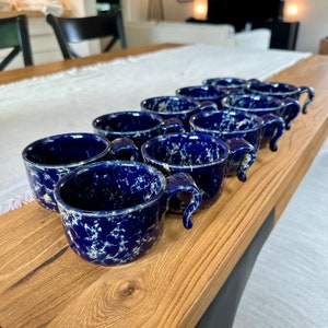Può includere: Un set di dieci tazze in ceramica blu e bianca con una glassa macchiettata. Le tazze hanno una forma arrotondata e un singolo manico.