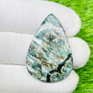 May include: A teardrop-shaped gemstone with a mix of green, brown, and black patterns. The stone has a polished surface and is held by a gloved hand. The gemstone's colors and patterns create a unique, natural appearance.