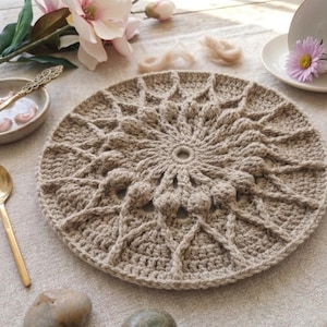 May include: A round, beige crocheted placemat with a detailed, textured design. The placemat is surrounded by a neutral-colored linen tablecloth. A gold spoon and a small bowl are visible.