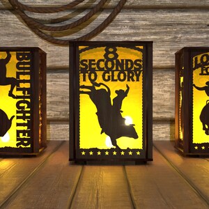 May include: Three wooden silhouette lanterns with a cowboy riding a bucking bull. The lanterns are lit and glow yellow. The first lantern says "Bullfighter" with three stars. The second lantern says "8 Seconds to Glory". The third lantern says "Looking for 8".